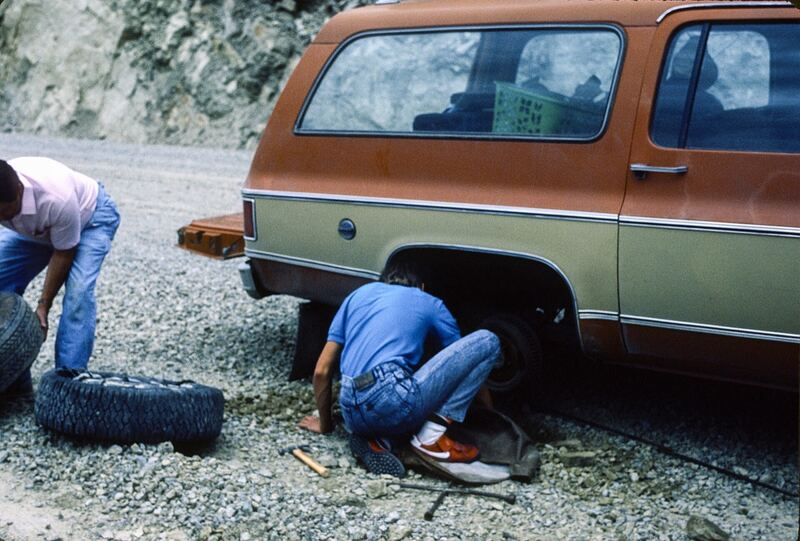 Two people changing a flat tire on an older SUV on a gravel roadside (similar vehicle)