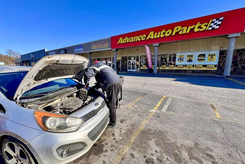 Auto parts store worker replacing a car battery under an open hood in a parking lot (similar vehicle)