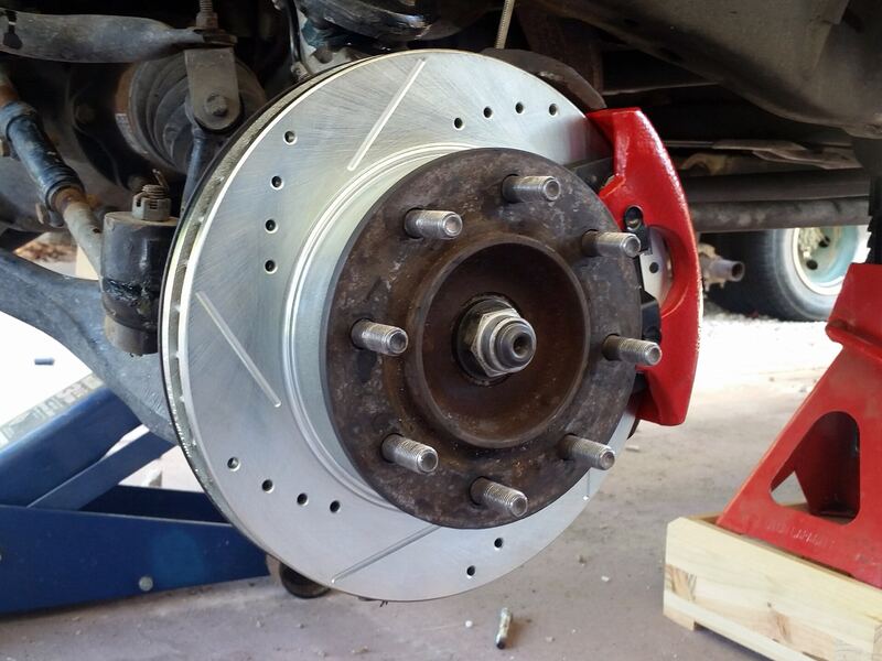 Wheel removed on a vehicle on jack stands, showing a new drilled brake rotor and red caliper (similar vehicle)